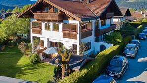 Um chalé de dois andares com telhado inclinado, varandas de madeira e um jardim com uma árvore e área de estar ao ar livre.