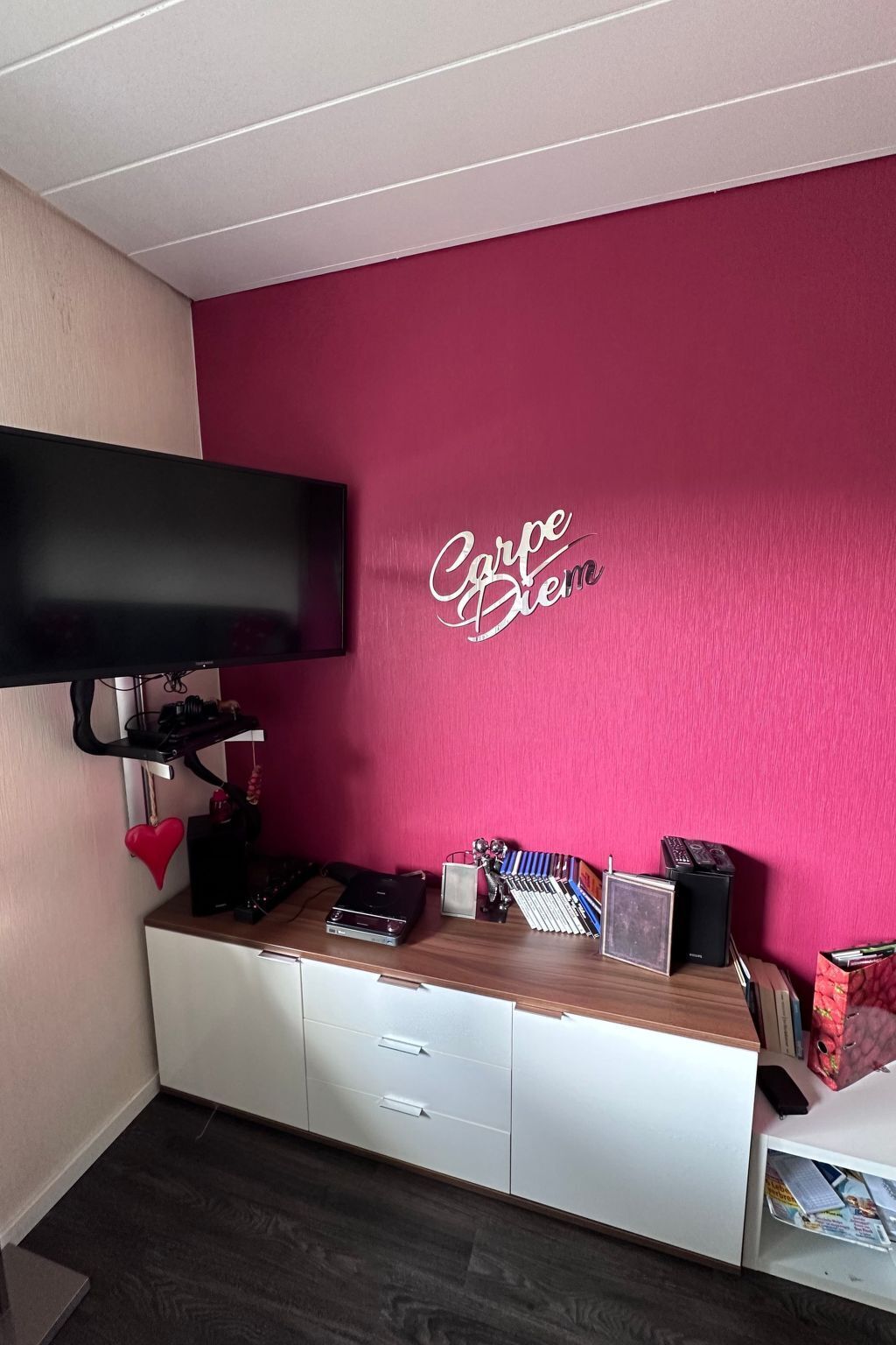 A room with a pink wall, a mounted TV, a white cabinet with drawers, and a shelf with various items.