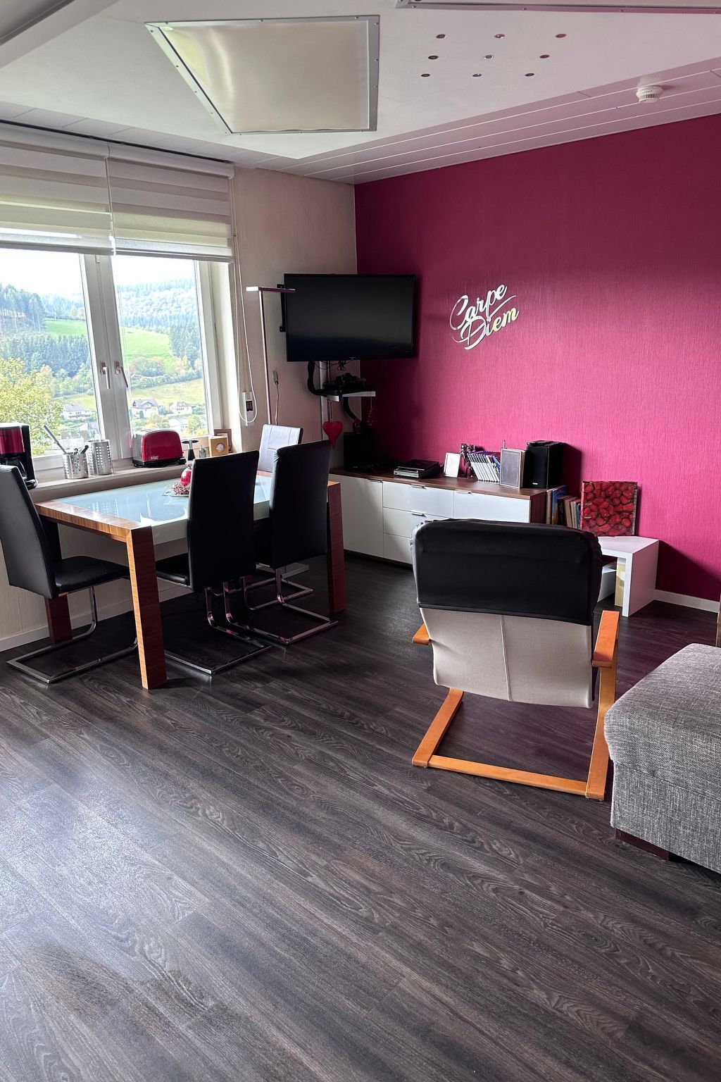 Un salon moderne comprenant un coin repas, une grande fenêtre offrant une belle vue, un téléviseur à écran plat et un mur d’accent rouge.