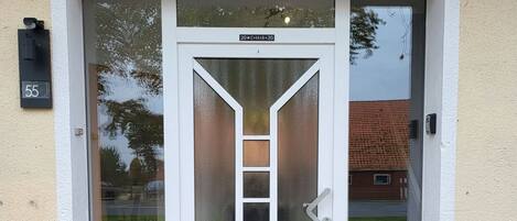 A building entrance with a glass door, a metal number plate, and a tiled staircase.