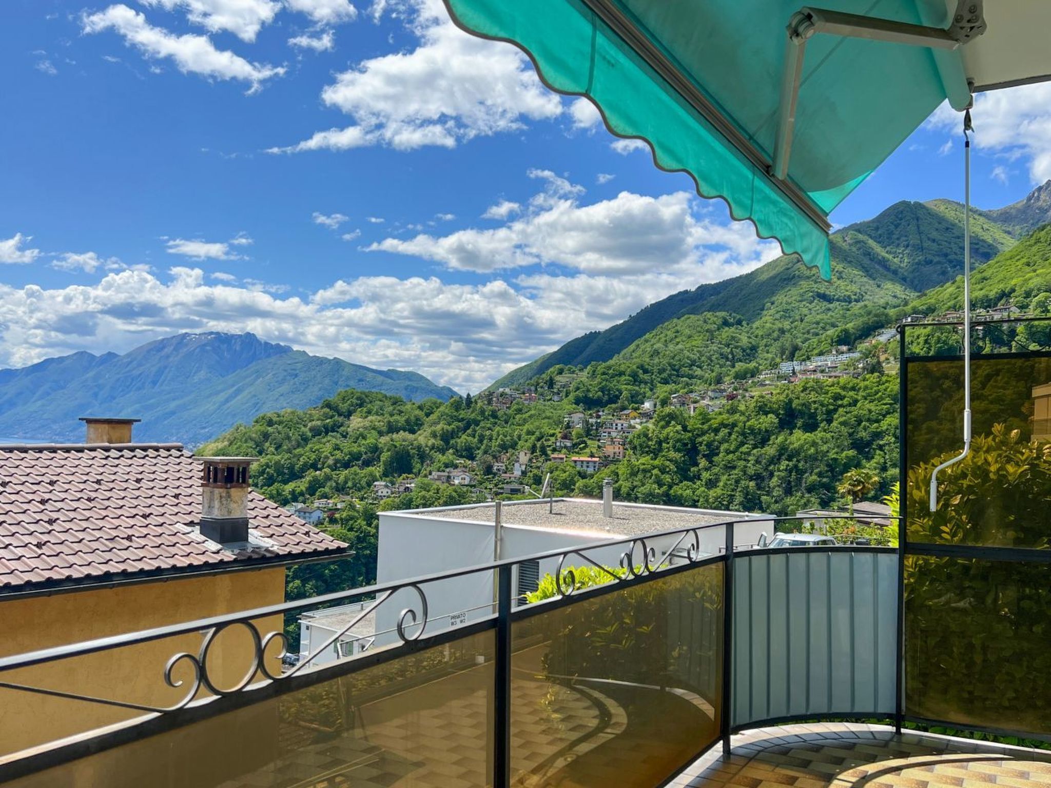 Ein Balkon mit Blick auf eine gebirgige Landschaft, ein Ziegeldach und ein grünes Sonnensegel.