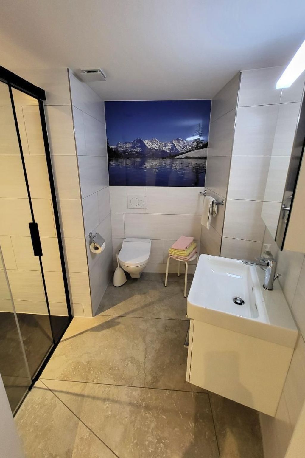 Pieni kylpyhuone, jossa on wc, pesuallas ja pieni jakkara. Suuri seinämaalaus esittää vuoristomaisemaa.