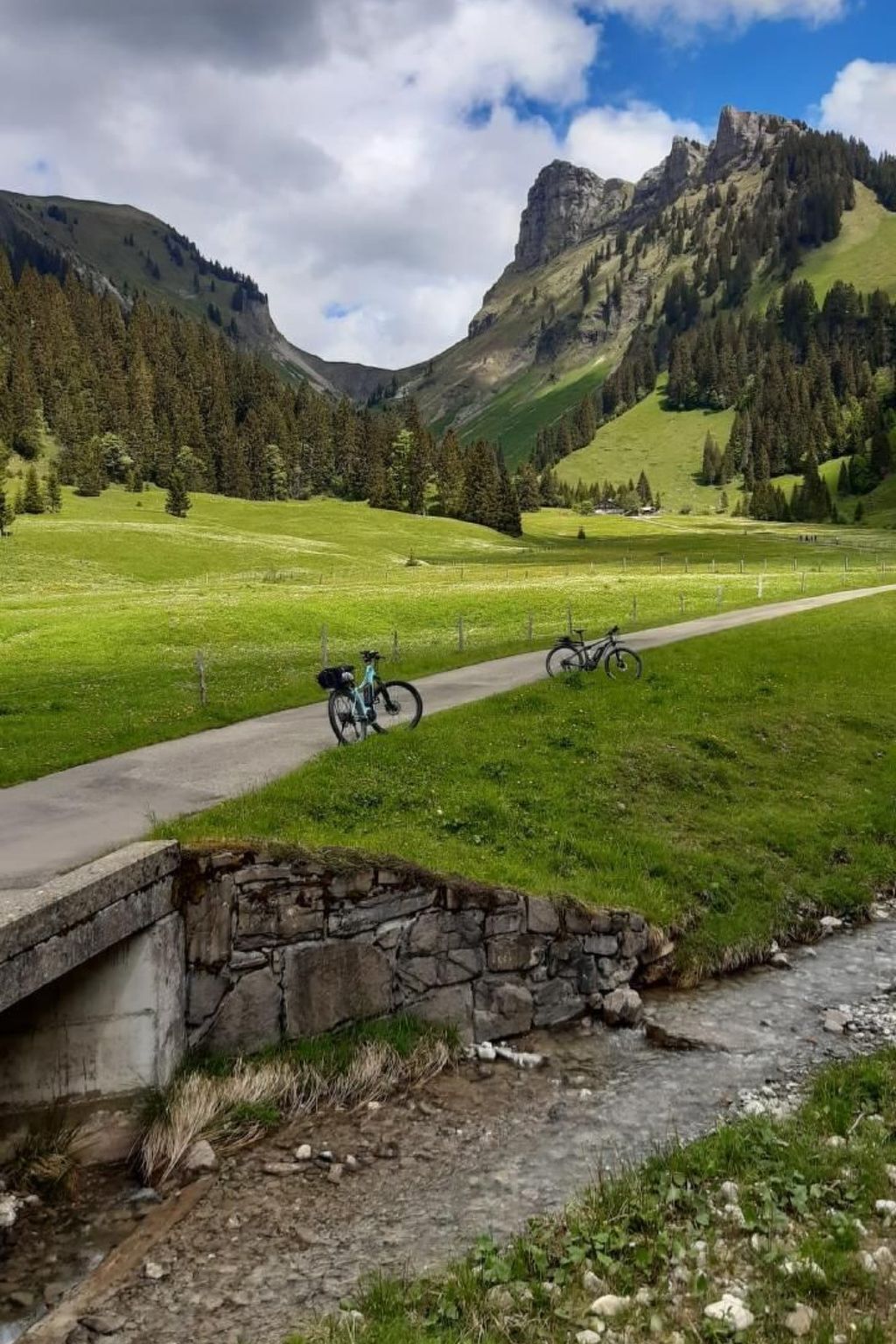 A stone bridge over a stream, a paved path, and bicycles parked along the path in a mountainous landscape.