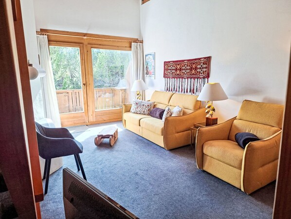 A living room with a sofa, armchair, and a small table. There is a window with curtains and a view of greenery outside.