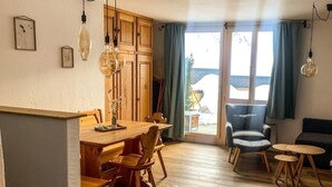 Een gezellige woonkamer met een houten eettafel, stoelen, een blauwe fauteuil en een bank. Er hangen lampen en er is uitzicht buiten door een glazen deur.