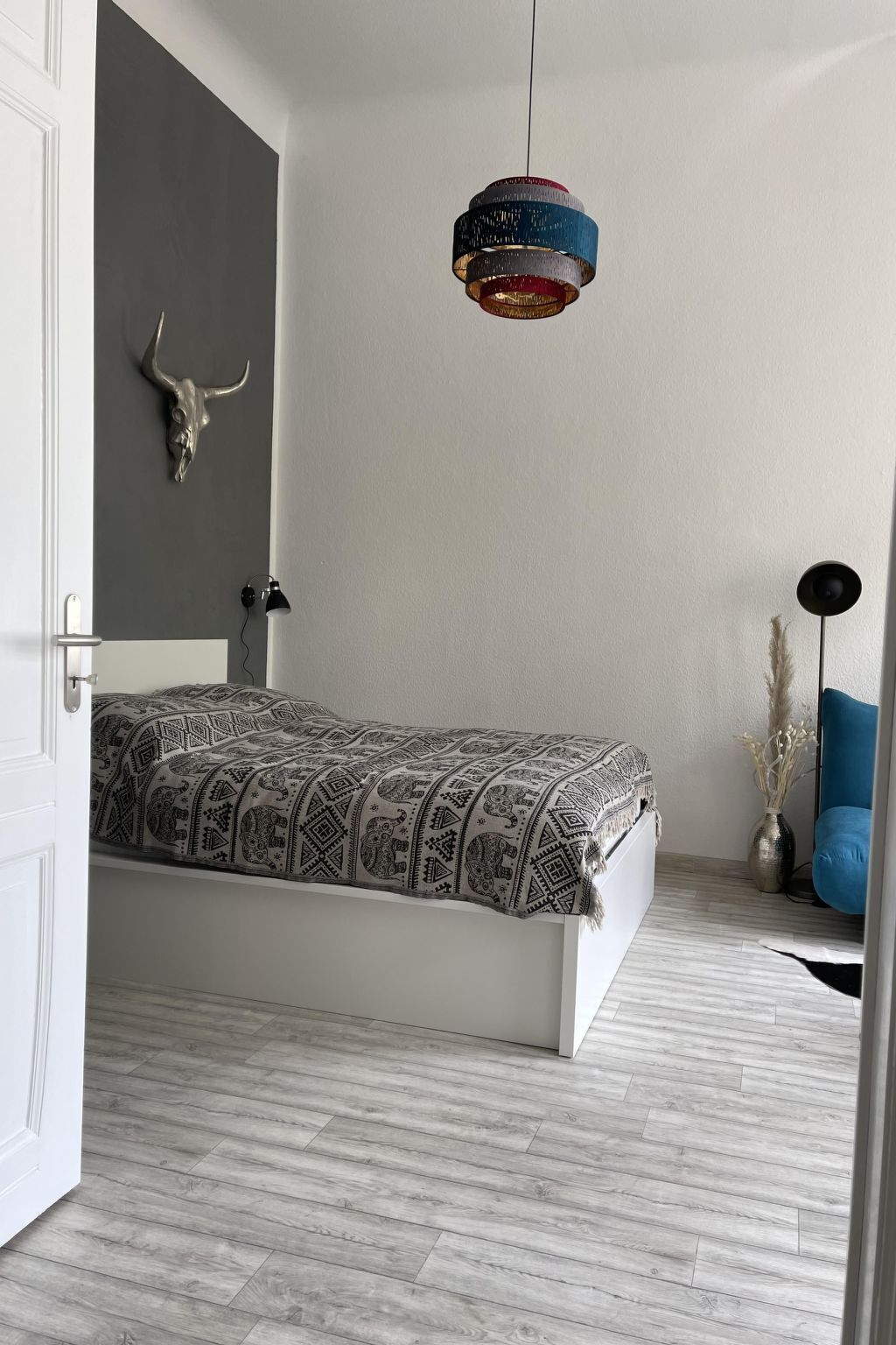 Ein schlichter Schlafzimmer mit Bett, einer Pendelleuchte, einer Wanddekoration in Form eines Schädels und einem kleinen Nachttisch mit einer Vase.