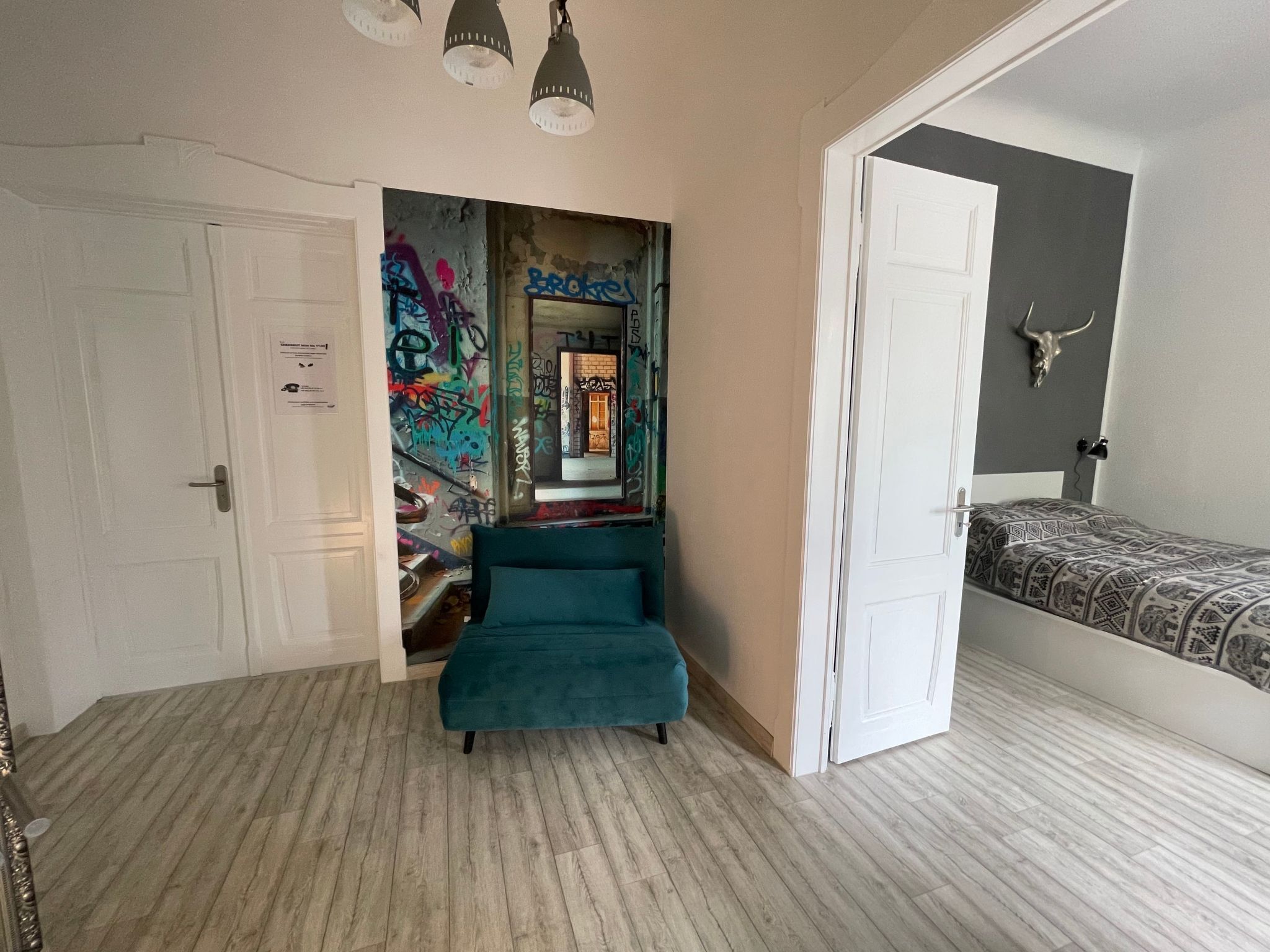 A room with a teal sofa, a bed with patterned bedding, a mirror with graffiti, and a door with a white frame.
