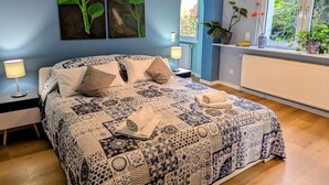 A bedroom with a patterned bedspread, two wall-mounted paintings of sunflowers, a window with a view of greenery, and a radiator.
