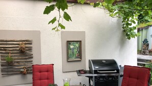 A patio with a round wooden table, red cushioned chairs, a barbecue grill, and potted plants on a wooden rack.