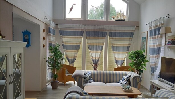 A spacious living room with a large sofa, a television, and a dining table. There are two potted plants and a clock on the wall. - Haus Bernstein by Interhome (Damshagen)
