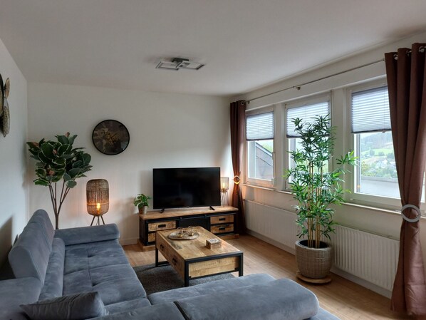 A living room with a grey sofa, a wooden coffee table, a flat-screen TV, and a large window with curtains.