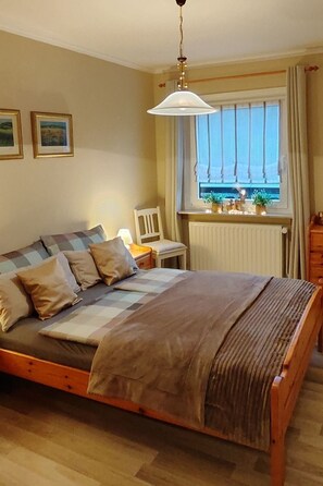 Une chambre à coucher avec un lit en bois, une fenêtre avec des rideaux, un radiateur et des tableaux encadrés accrochés au mur.