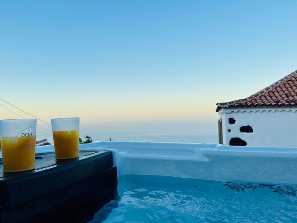 A poolside view with two glasses of orange juice on a black surface, overlooking a body of water and a building with a tiled roof.