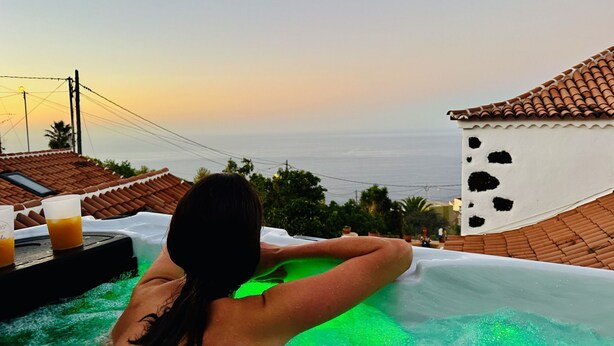 Una persona relajándose en un jacuzzi con vista al mar y un edificio con techo de tejas.
