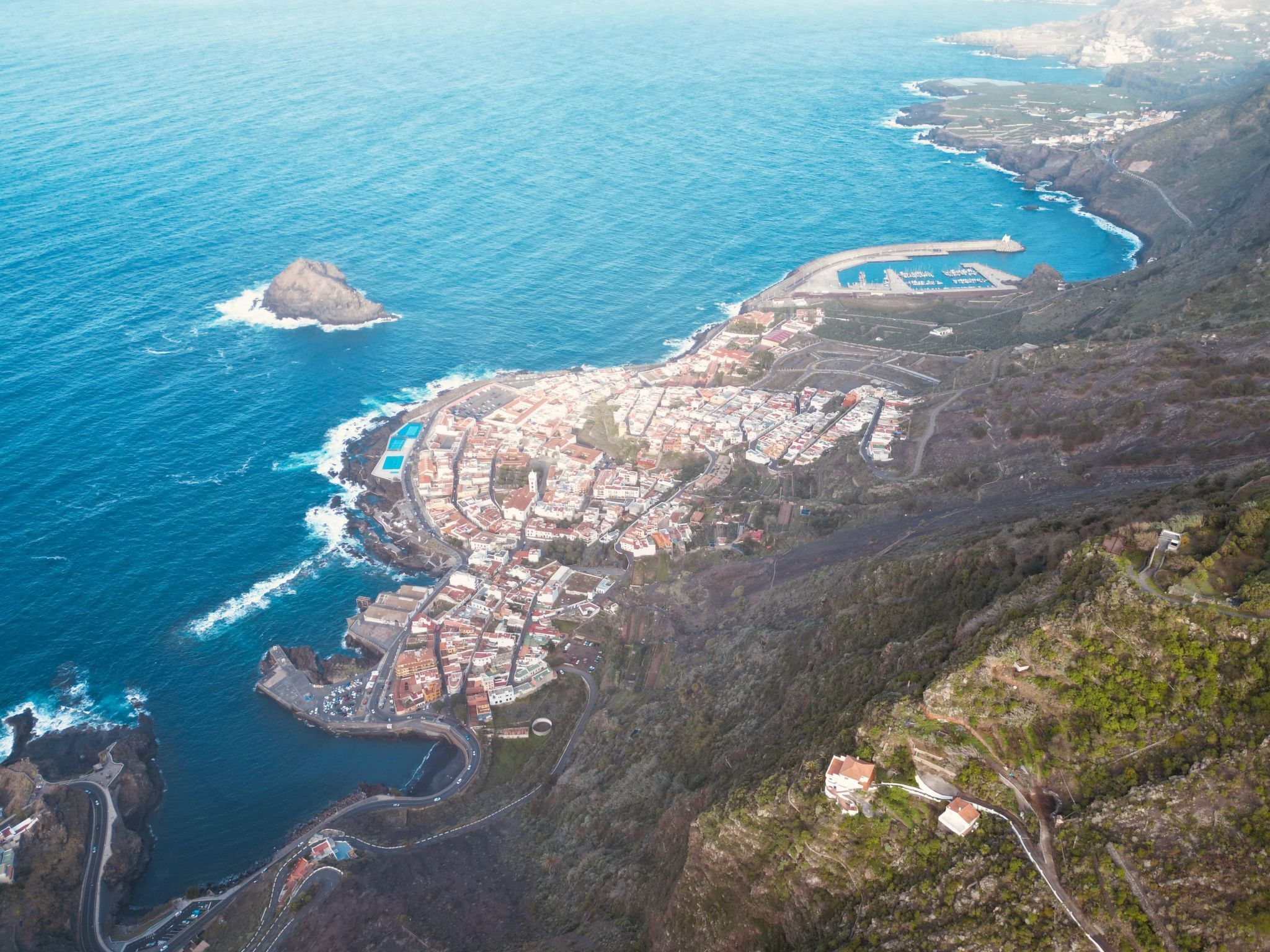 Vista aérea de un pueblo costero con un conjunto central de edificios, un puerto deportivo y una costa escarpada.
