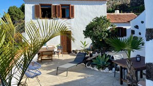 Un edificio blanco con techo de tejas y contraventanas de madera, rodeado de un patio con mesa, sillas y plantas.