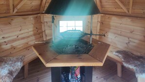 A wooden cabin with a central wood-burning stove, a window, and a bench.