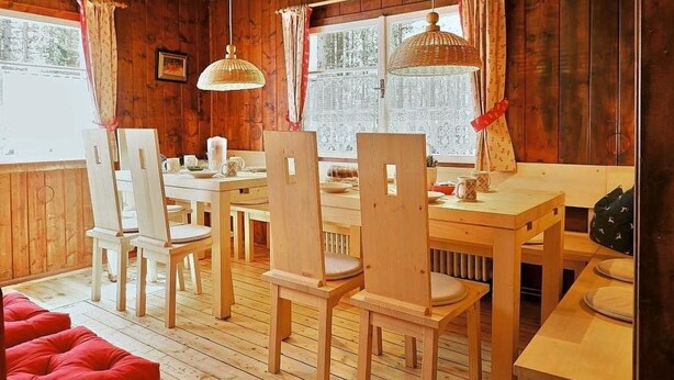 A wooden cabin with a dining area featuring a wooden table and chairs, a hanging light fixture, and a view through the window. - Berg Hütte Hochpillberg by Interhome (Pill)