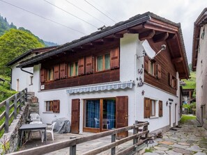 Un chalet de dos pisos con contraventanas de madera, una escalera de piedra y una pequeña zona de estar al aire libre.