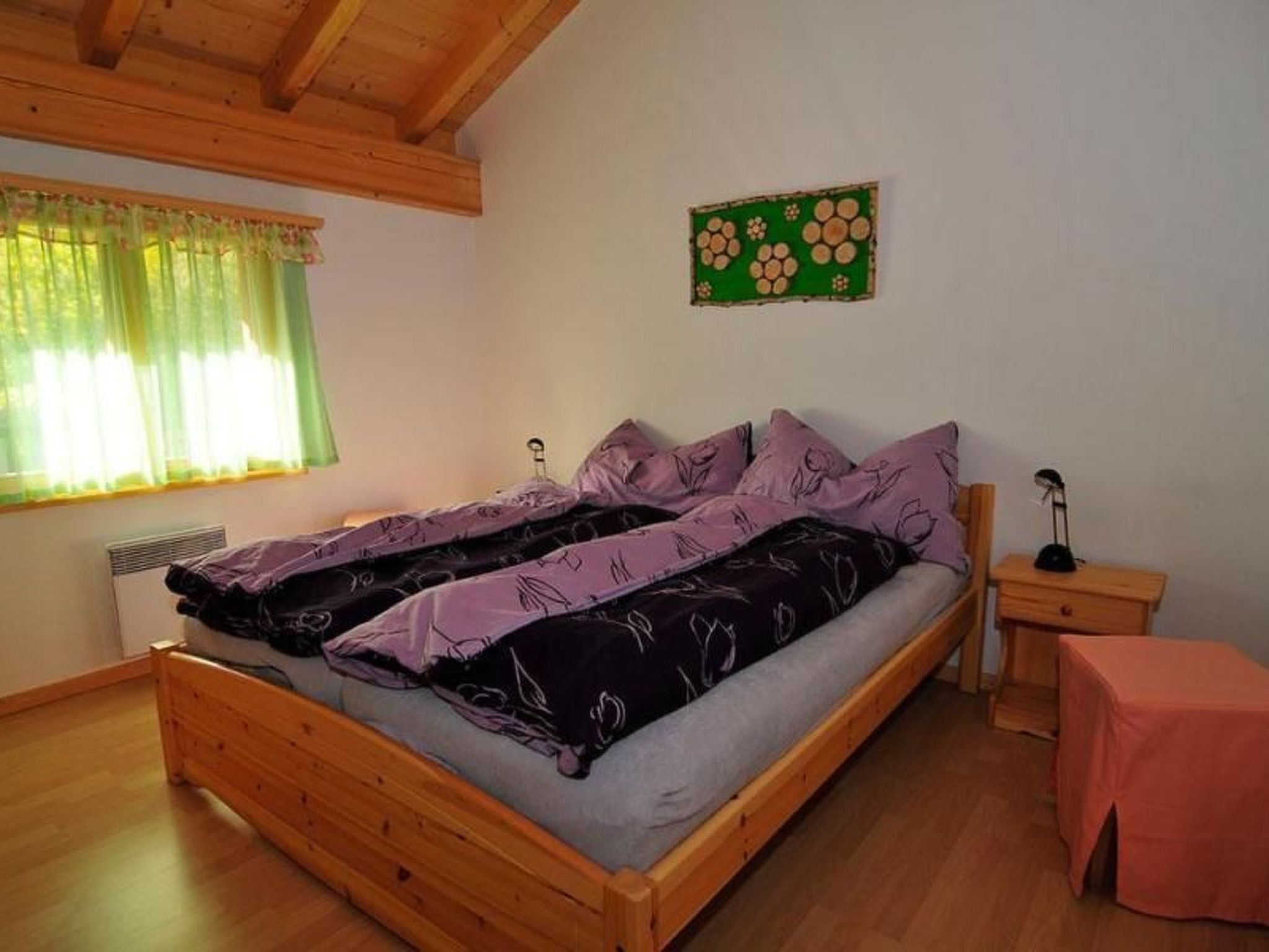 Une chambre à coucher avec un lit en bois, une table de chevet, une table, une fenêtre avec des rideaux et un élément décoratif mural.