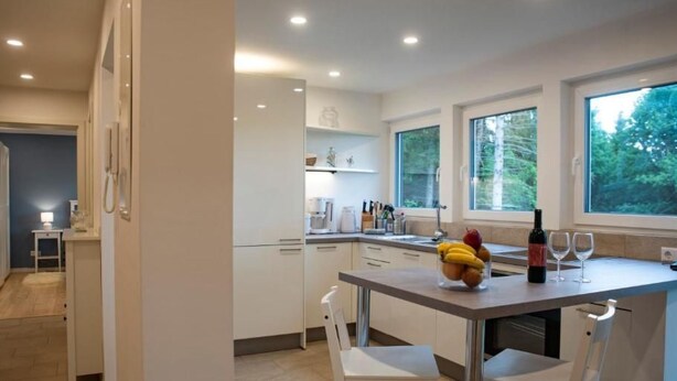A modern kitchen with white cabinets, a dining table with two chairs, a fruit bowl, a wine bottle, and wine glasses. - Fewo Am Sommerberg by Interhome (Donaueschingen)