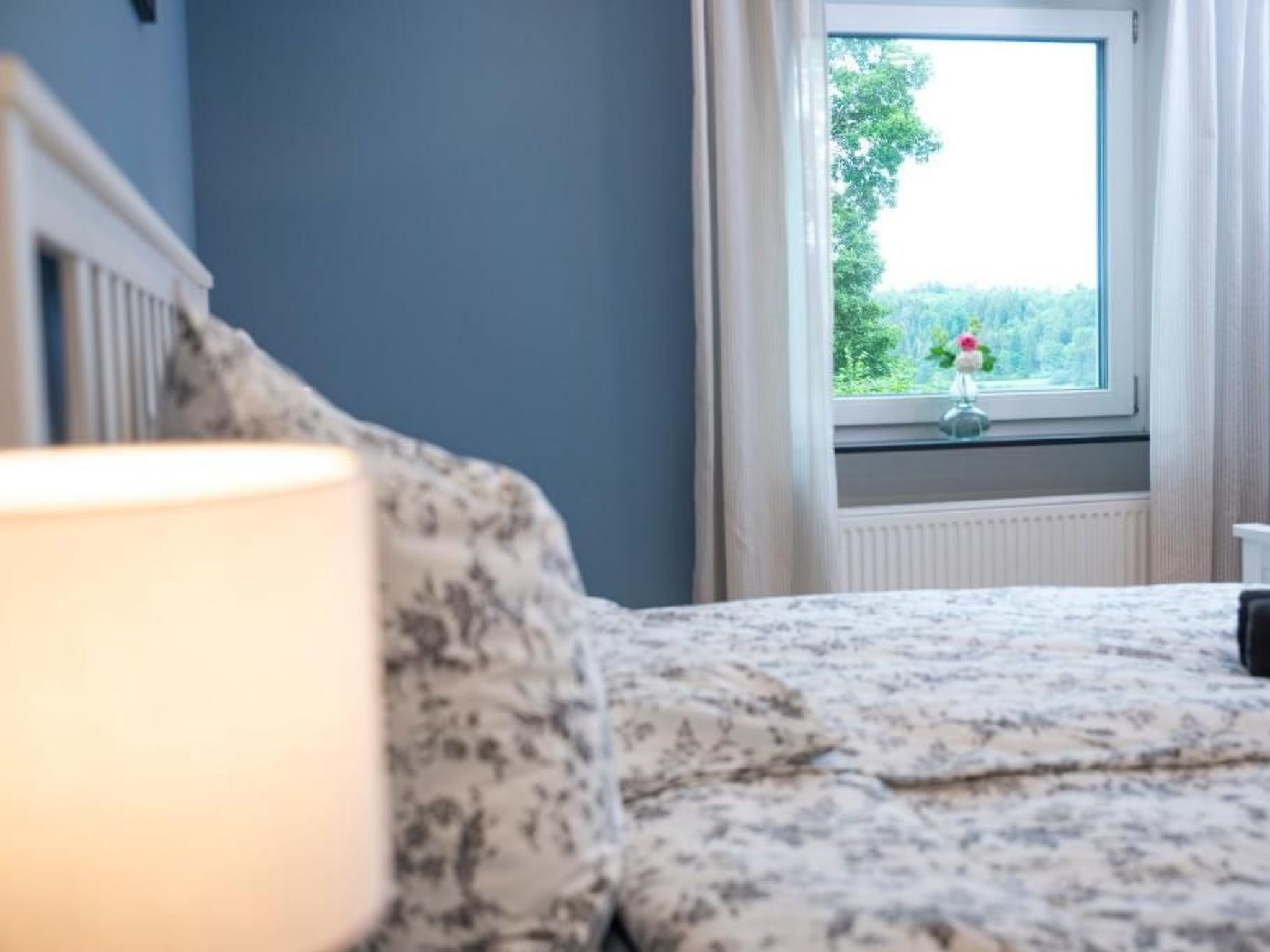 Een slaapkamer met een bed, een raam met uitzicht op groen en een nachtkastje met een lamp.