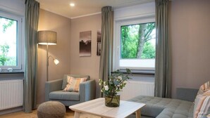 A modern living room with a grey sofa, a blue armchair, a wooden coffee table, and a large window with curtains. - Fewo Am Sommerberg by Interhome (Donaueschingen)