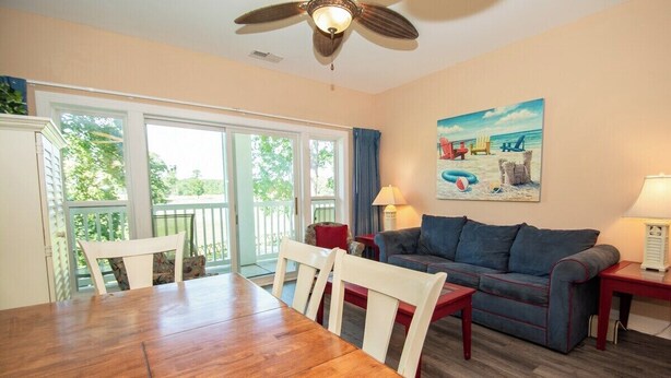 A living room with a wooden dining table, white chairs, a blue sofa, and a ceiling fan. - World Tour 104-202a (Myrtle Beach)