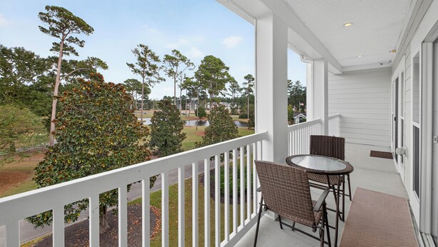 A balcony with wicker chairs and a table, overlooking a grassy area with trees and a body of water. - World Tour 1009-302 (Myrtle Beach)