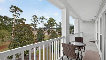 A balcony with wicker chairs and a table, overlooking a grassy area with trees and a body of water.