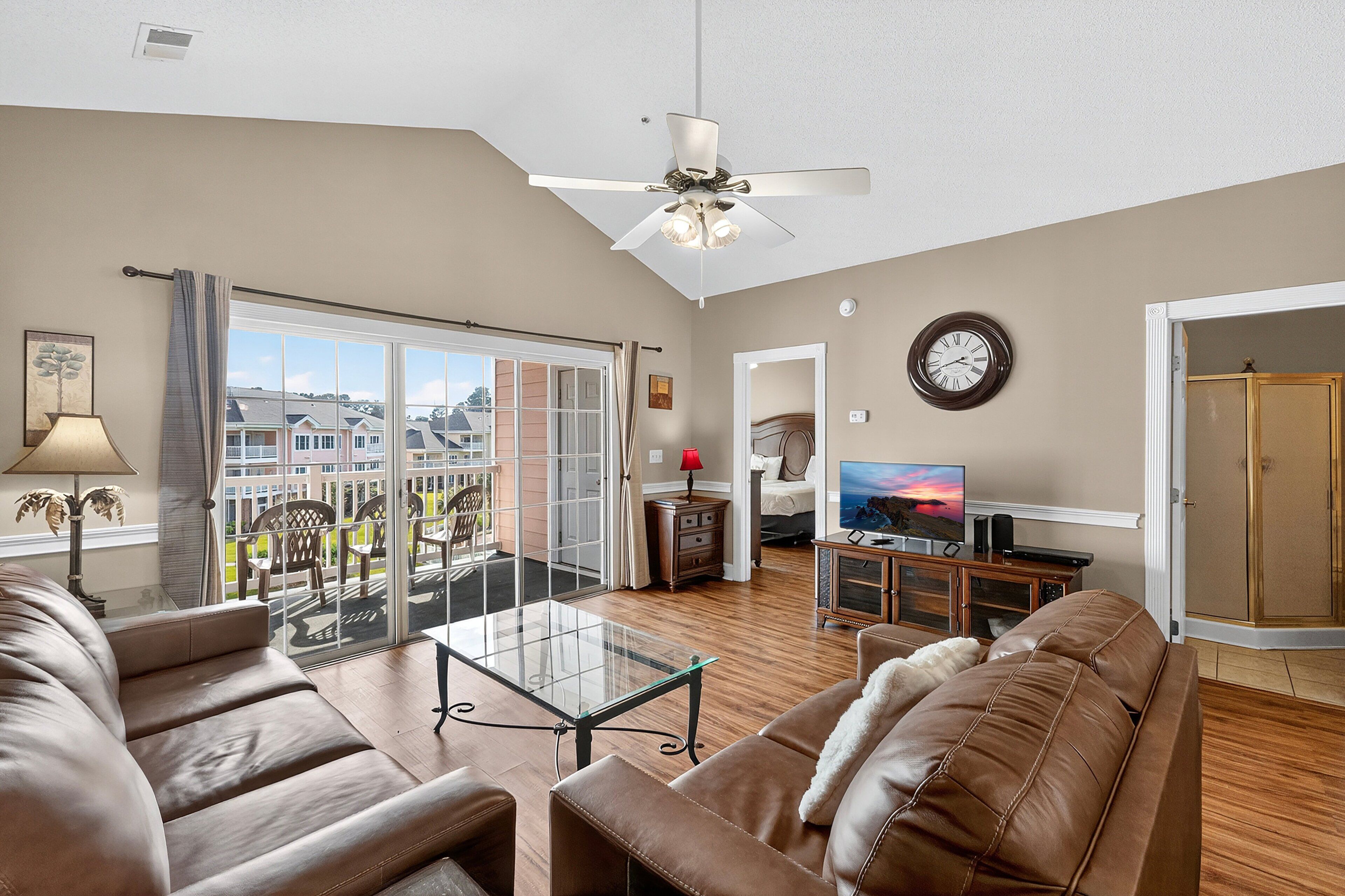 A living room with a glass coffee table, leather sofas, a TV, and a balcony with a view of houses.