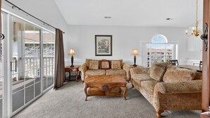 A spacious living room with a large sliding glass door leading to a balcony, a wooden coffee table, patterned sofas, and a chandelier. - Magnolia Place 4687-304 (Myrtle Beach)