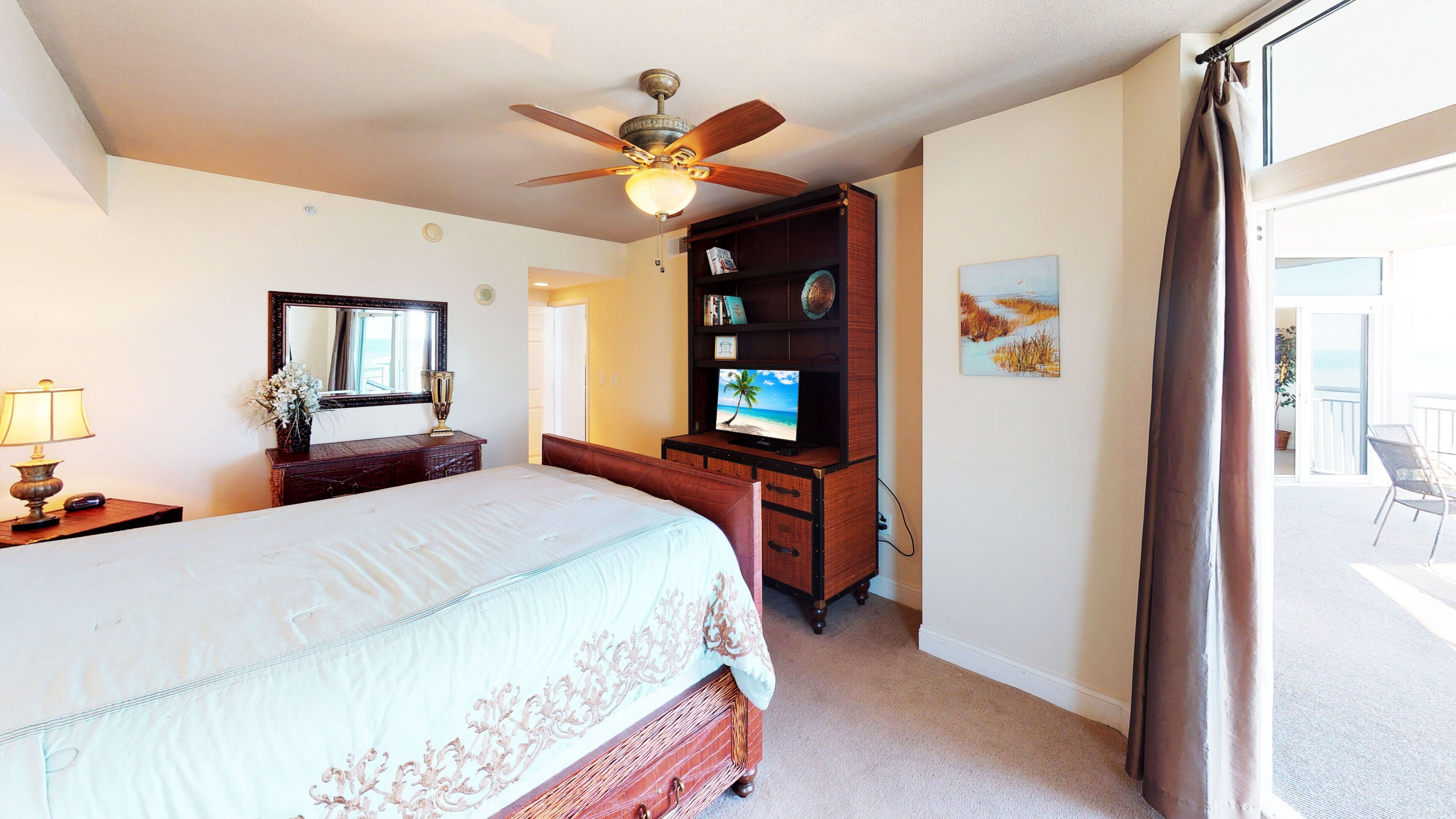 A hotel room with a bed, a desk with a television, a ceiling fan, a mirror, and a view of the outdoors.