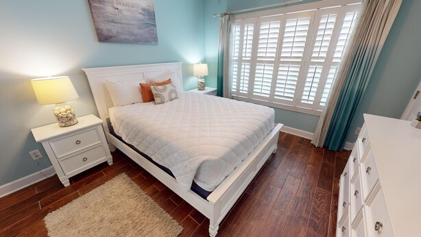 A bedroom with a white bed, two bedside tables with lamps, a nightstand, a dresser, a window with shutters, and a painting on the wall. - Magnolia North Unit 105 (Myrtle Beach)