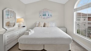 Quarto com uma cama grande, cômoda, mesa de cabeceira, abajur e janela com vista para edifícios.