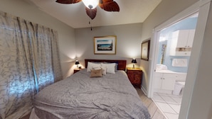 Una camera da letto con un letto, comodini, una ventola a soffitto, un quadro e un bagno visibile attraverso una porta aperta.