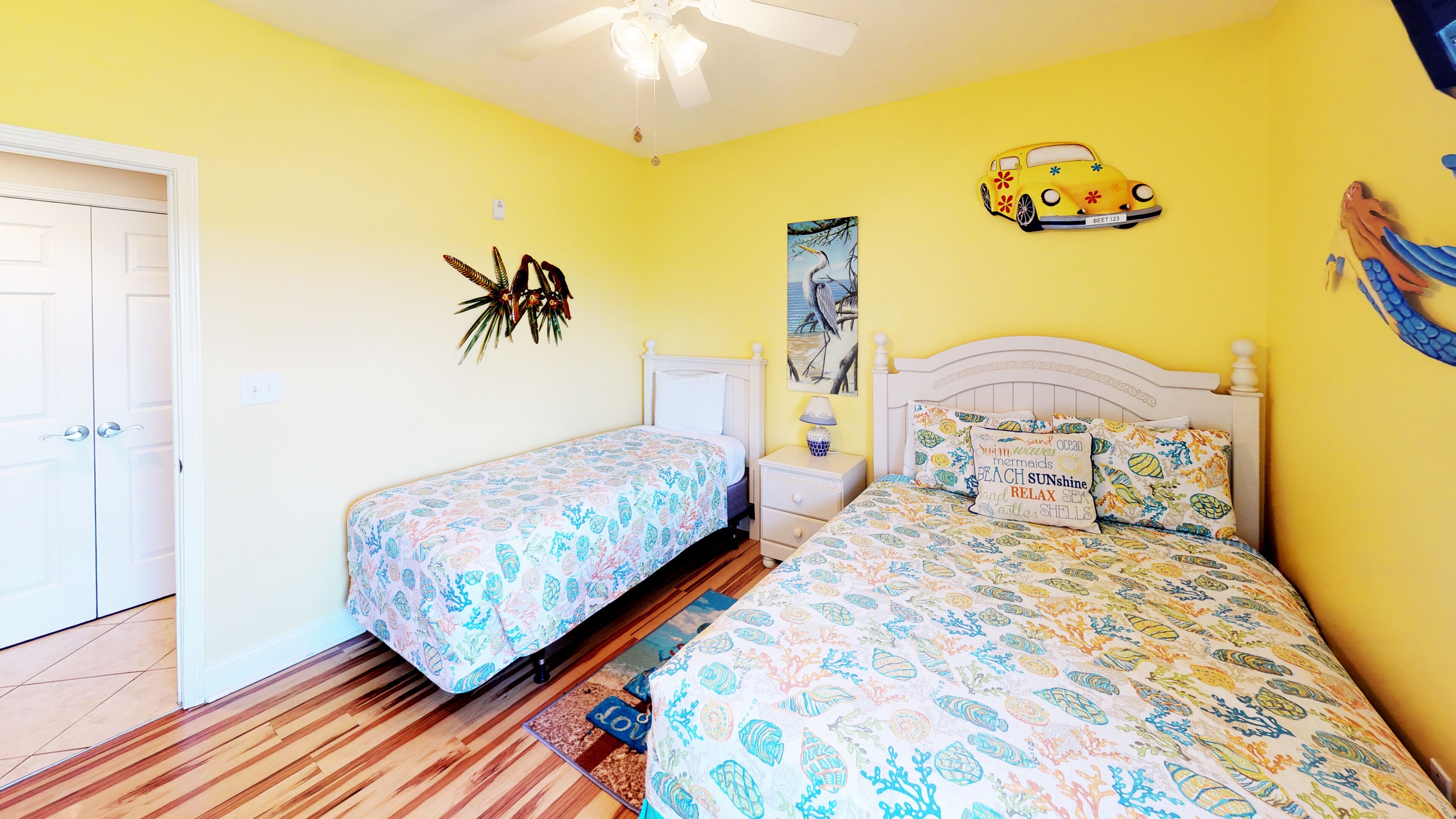 A bedroom with two beds, a ceiling fan, a door, and a wall with a car and a bird decoration.