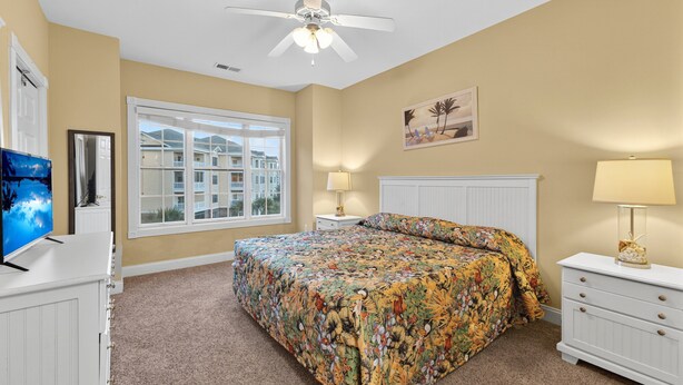 A bedroom with a large bed, a ceiling fan, a window with a view of buildings, and a TV on a dresser. - Magnolia Pointe Luster Leaf 4821-301 (Myrtle Beach)