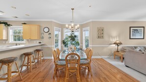 A spacious dining area with a glass-top table surrounded by wicker chairs, a chandelier, and a view of a green plant through the windows. - Magnolia Pointe Luster Leaf 4821-301 (Myrtle Beach)