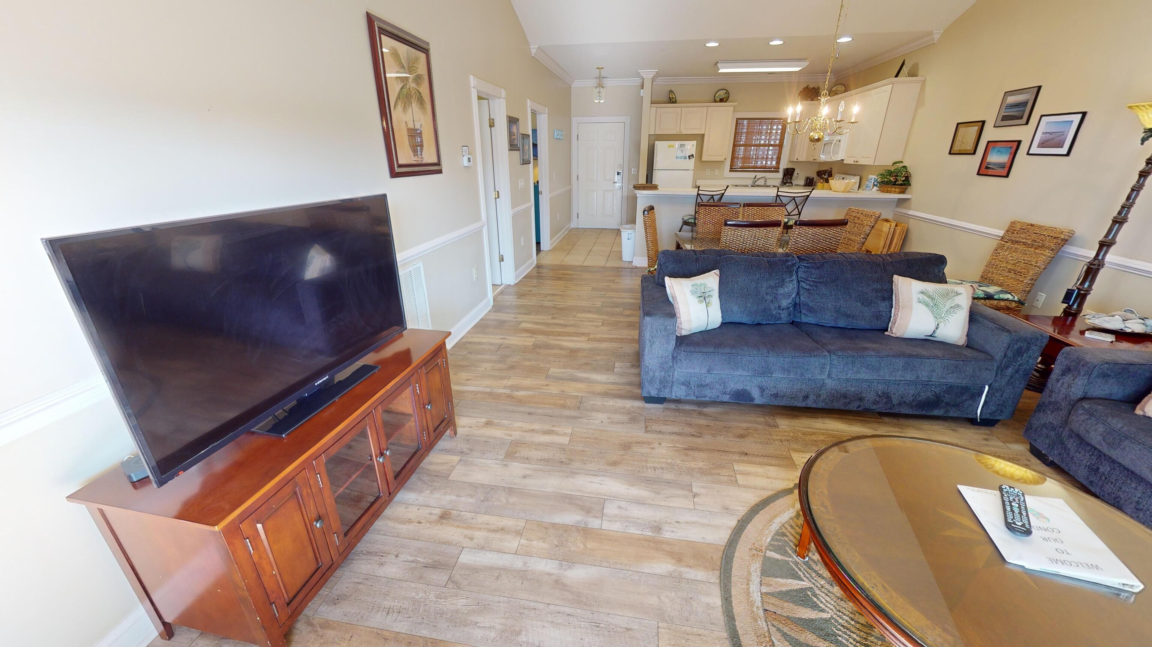 Un soggiorno con una TV a schermo piatto su un mobile in legno, un divano blu con cuscini decorativi, un tavolino rotondo e, sullo sfondo, una cucina con pensili in legno e un lampadario.