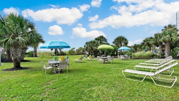 A grassy outdoor area with lounge chairs and umbrellas, surrounded by palm trees and a clear sky. - Meridian Plaza 1401 (Myrtle Beach)