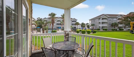 A covered outdoor area with a round table and chairs, a white fence, and a view of a building complex with multiple balconies and a well-maintained lawn.