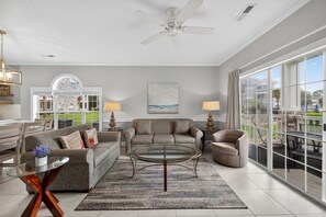 A spacious living room with a large glass coffee table, comfortable seating, and a view of the outdoors through large windows. - Magnolia Place 4695-102 (Myrtle Beach)
