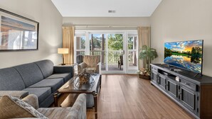 A living room with a grey sofa, a coffee table, a TV on a cabinet, and a view of the outdoors through large glass doors. - World Tour 104-303 (Myrtle Beach)