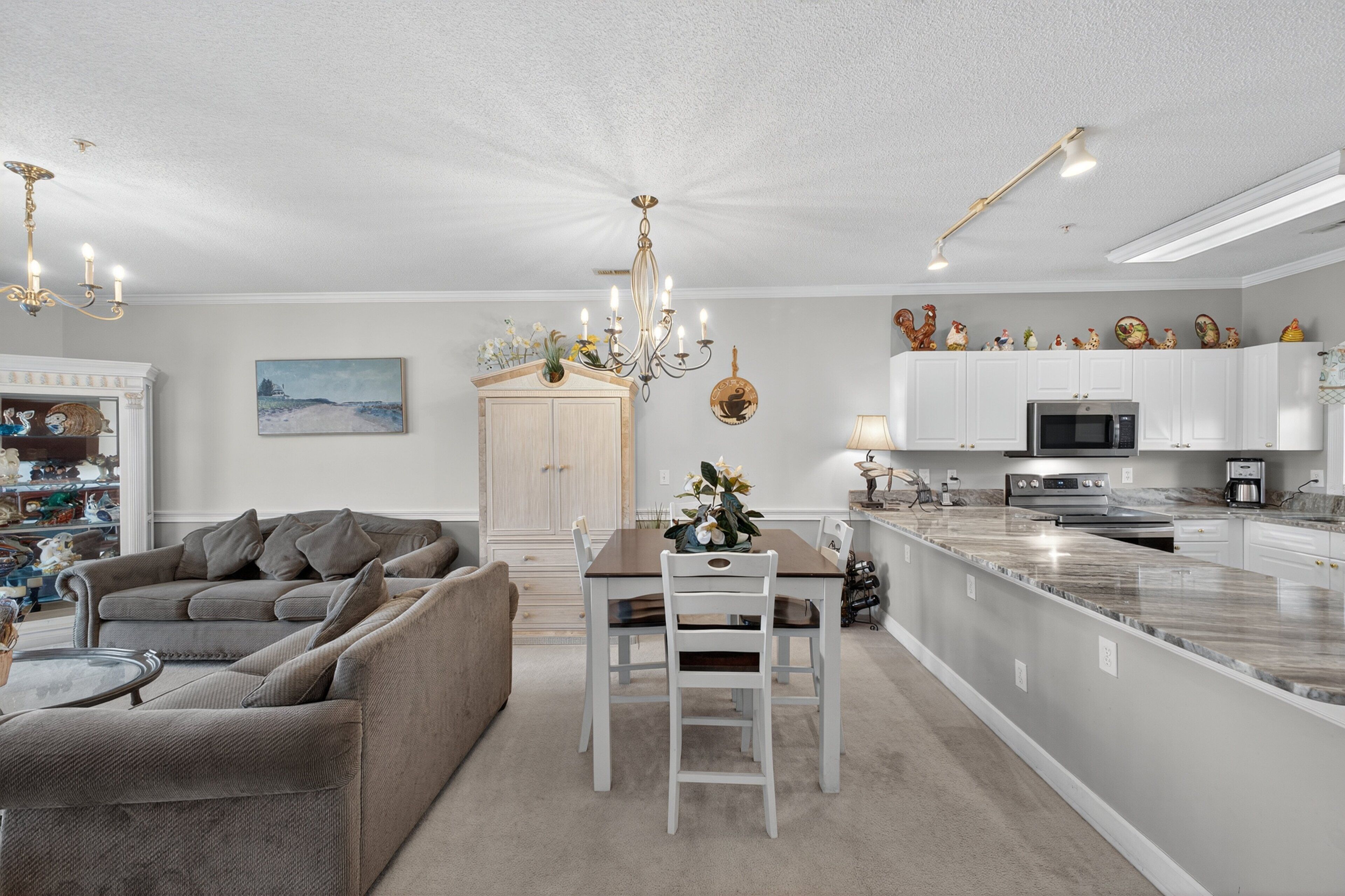 Ein modernes Wohnzimmer mit grauem Sofa, einem Esstisch mit Stühlen, einer Chandelierleuchtre und einer Küchenzeile mit weißen Schränken und Geräten.