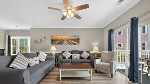 A living room with a grey sofa, a wooden coffee table, a grey armchair, a ceiling fan, and a large window with curtains. - Magnolia North 4827-201 (Myrtle Beach)