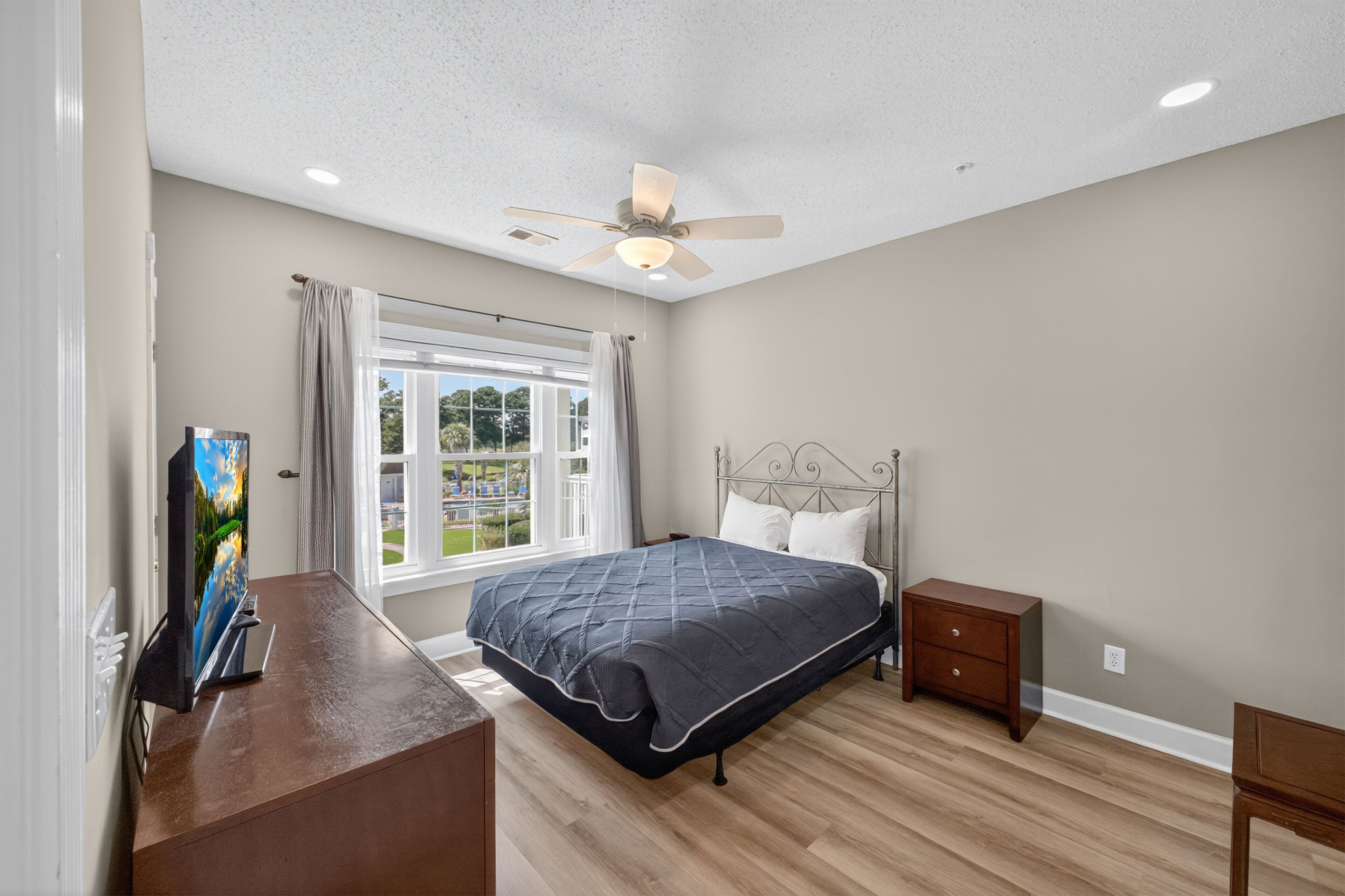 Une chambre à coucher comprenant un lit, une table de chevet, une commode, un ventilateur de plafond et une fenêtre avec des rideaux.