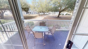 Une terrasse extérieure couverte, avec une table et des chaises, une balustrade blanche et une vue sur un espace aménagé avec des arbres et un bâtiment en arrière-plan.