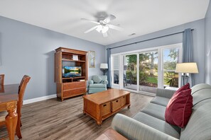 Una sala de estar con una mesa de centro de madera, un sofá gris, un mueble de entretenimiento de madera y un gran ventanal corredizo que da a un patio exterior.