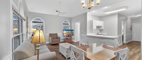 A spacious living room with a sofa, a dining table, and chairs, a kitchen area, and a large window with a view.